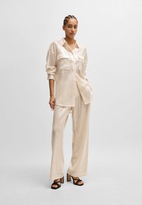 Cremfarbens, paillettenbesetztes Shirt und weit geschnittene Hosen mit zwei Brusttaschen. Kombiniert mit schwarzen Blockabsatzsandalen. Glatte, glänzende Textur.