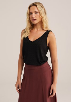 Donna bionda che indossa una canotta nera senza maniche con texture e una gonna marrone a vita alta, in piedi contro uno sfondo chiaro uniforme.