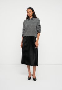 Donna in piedi, che indossa un maglione grigio sopra una camicia a righe nera, una gonna midi plissettata nera e tacchi a punta neri, su uno sfondo neutro.