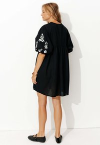 Robe noire avec des manches bouffantes, broderie florale blanche sur les manches, coupe ample, longueur genou, associée à des ballerines noires à clous.