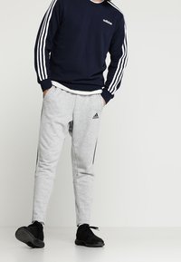 Sudadera navy con logo blanco de Adidas y tres bandas blancas en las mangas, combinada con joggers gris claro que tienen franjas negras a los lados.