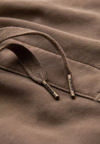 Détail en tissu marron avec une texture tissée ; deux boucles en tissu marron avec des étiquettes métalliques portant les inscriptions "DIDBARE" et "PUMBARE."