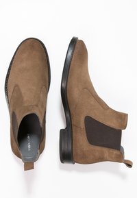 Botas Chelsea de ante marrón con paneles laterales elásticos, suelas de goma negra y un diseño suave y estructurado. Acabado mate con acentos mínimos.