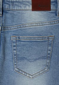 Jeans de mezclilla azul claro con un bolsillo trasero con costuras naranjas y una etiqueta de cuero marrón en la cintura, embosada con el nombre de la marca.
