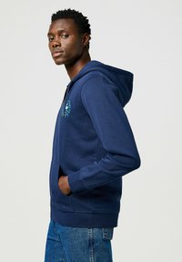 Felpa con zip color blu navy, cappuccio regolabile con lacci, tasca kangaroo e dettagli del logo ricamati sul petto. Tessuto morbido e texturizzato.