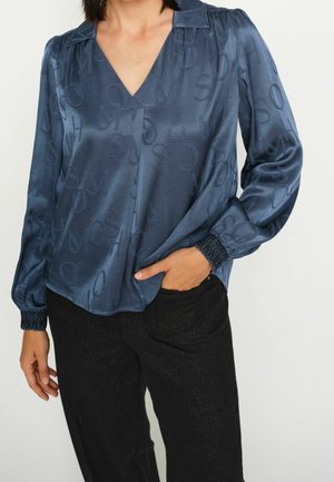 Femme porte un chemisier bleu foncé à manches longues avec des lettres texturées et un pantalon noir, main partiellement dans la poche, sur un fond uni.