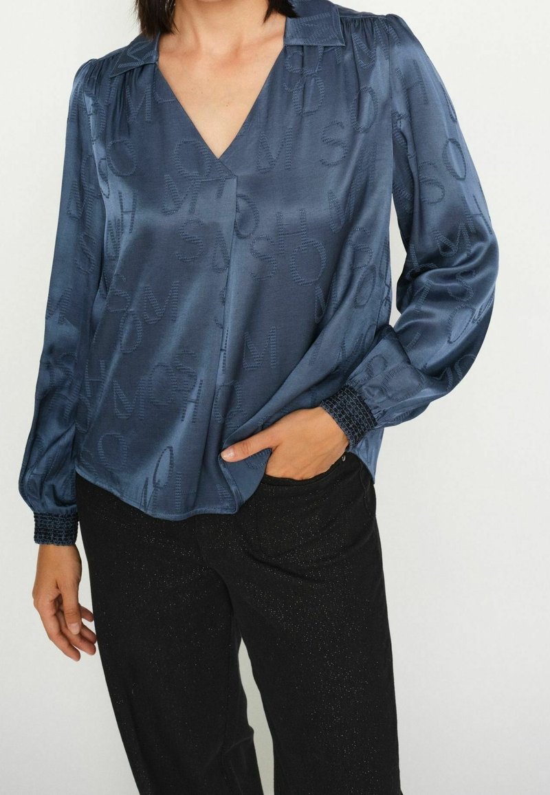 Femme porte un chemisier bleu foncé à manches longues avec des lettres texturées et un pantalon noir, main partiellement dans la poche, sur un fond uni.