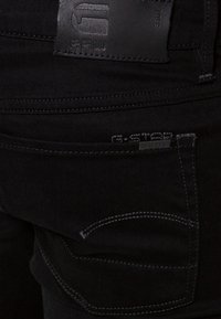 Mustat denim-farkut, joissa on nahkapatentti kohokuvioidulla logolla, kontrastiväriset ompeleet ja takataskudellinen yksityiskohta.