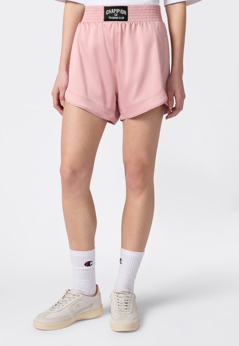 Personne portant un short à taille élastique rose marqué "Champion Training Club", des chaussettes blanches mi-mollet et des baskets beige, debout sur fond blanc.