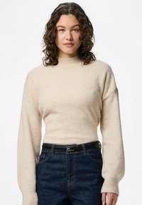 Femme aux cheveux bouclés portant un pull à col roulé beige et un jean bleu foncé avec une ceinture noire, debout devant un fond blanc uni.