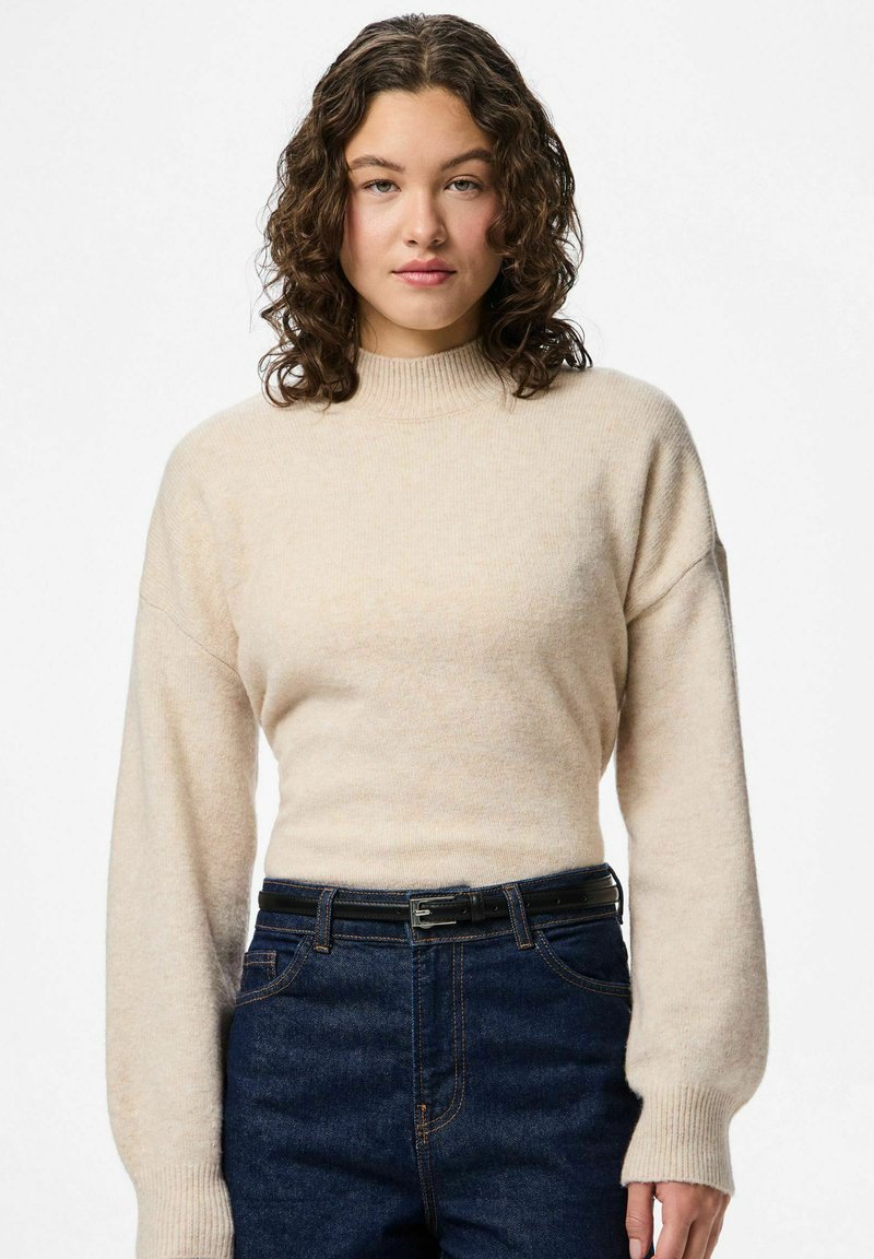 Femme aux cheveux bouclés portant un pull à col roulé beige et un jean bleu foncé avec une ceinture noire, debout devant un fond blanc uni.
