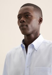 Camicia blu chiaro con bottoni, realizzata in un tessuto testurizzato, colletto a pieghe e taschino sul lato sinistro del petto. Il colletto interno presenta un accento con motivo.