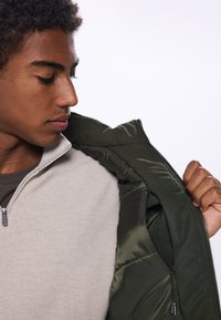 Light gray zip-up sweatshirt with fine texture, layered under a dark green padded jacket. Zipper detail visible, showcasing the inner lining.