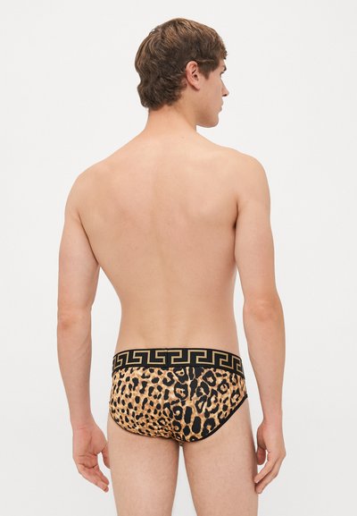 Junger Mann steht mit dem Rücken zur Kamera und trägt einen Leopardenmuster-Slip mit einem schwarz-goldenen geometrischen Bund.