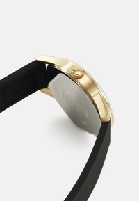 Gold-tone watch with a smooth case and black silicone strap. Features a textured crown, round shape, and a metal case back.