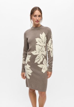 Femme portant une robe en maille taupe avec de grands motifs floraux blancs, manches longues et col rond, debout devant un fond blanc uni.