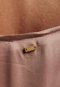 Bronzefarvet stofmærke med teksten "SUPERDRY STUDIOS" fastgjort til glat, lys taupe teksturstof, med en ren syningkant synlig.