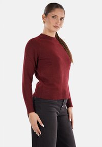 Maglione rosso a maglia con collo alto e maniche lunghe, caratterizzato da un design leggermente aderente e un tessuto testurizzato. Abbinato a jeans neri.