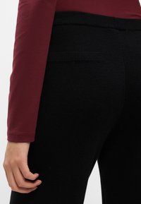 Mustad püksid tekstuuriga viimistlusega, millel on sobituv vöökoht ja väike tagakott. Külgedelt on nähtav burgundi värvi pika varrukaga topp.