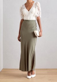 Olivgrön satinmaxiklänning med en sidoklyvning, parat med en vit spetsjacka med puffärmar. Accessoarer med en liten vit kuvertväska.