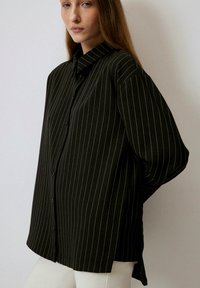 Black shirt with vertical white stripes, button front, collar, and loose fit. Long sleeves with dropped shoulders. Smooth fabric texture.