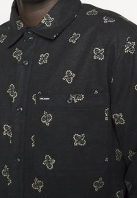 Zwart overhemd met knopen gemaakt van zachte stof met een crèmekleurig paisleypatroon, voorzien van een borstzak met logo en zwarte knopen.