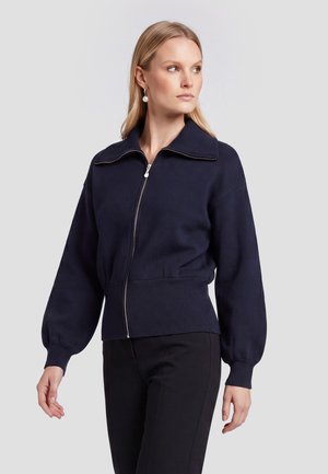 Lyshåret kvinde iført perleøreringe og en navy lynlås-sweater med ballonærmer, der ser mod højre mod en ensfarvet baggrund.