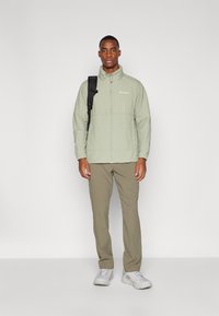 Light green waterproof jacket with a zip front, side pockets, and a high collar. Worn over beige pants and paired with gray sneakers.