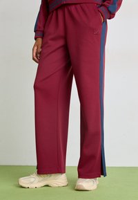 Bordeauxfarvede sweatpants med blå striber i siderne, lavet af blødt stof. Har en elastisk talje og slids ved anklerne. Samlet med beige sneakers.