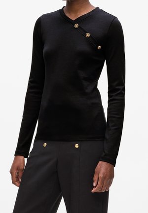 Haut noir ajusté à manches longues avec col en V et trois boutons dorés décoratifs sur une bande diagonale, associé à un pantalon noir avec deux boutons dorés.