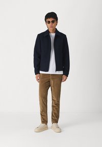 Giacca zip navy sopra una t-shirt bianca, abbinata a pantaloni in velluto a coste marroni e sneakers color crema. Gli accessori includono occhiali da sole rotondi.