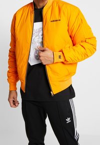 Orange vadderad bomberjacka med en svart Adidas-logotyp, ribbade muddkanter och dragkedja fram, bärs över en svart grafisk t-shirt och svarta byxor.