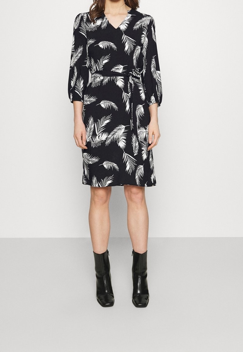 Femme portant une robe portefeuille noire mi-genou avec un motif de feuilles blanches, manches trois-quarts, et des bottines noires, sur fond uni.