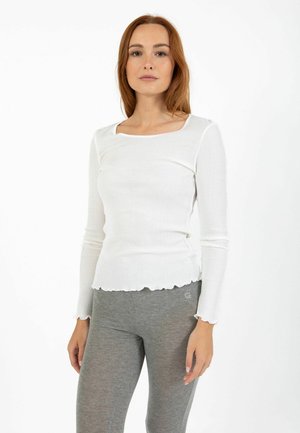 Woman with straight red hair wears a white long-sleeve ribbed top with lettuce hems and gray fitted pants against a white background.