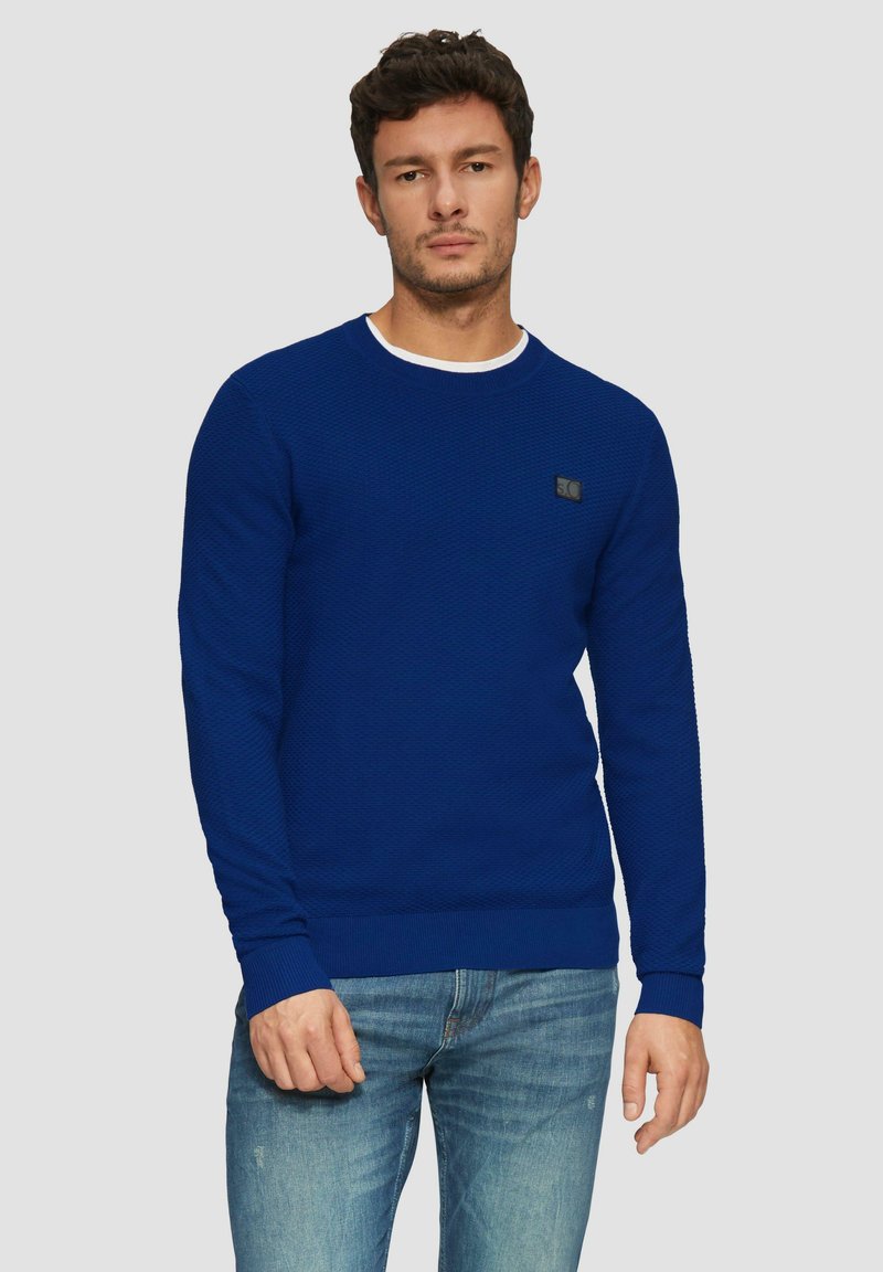 Blauer, gestrickter Rundhalsausschnitt-Pullover mit einem strukturierten Muster, gerippten Bündchen und Saum, ausgestattet mit einem kleinen schwarzen Logo auf der Brust. Getragen mit hellblauen Jeans.