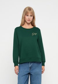 Sudadera verde hecha de una mezcla de algodón con puños y dobladillo de canalé. Presenta el logotipo dorado de "Gap" en el lado izquierdo del pecho. Combinada con jeans de color azul claro.