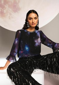 Blusa nera trasparente con stampa galattica, scollatura rotonda adornata da perline, abbinata a pantaloni frangiati neri con paillettes. Sullo sfondo si distingue una grande luna.