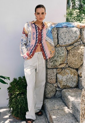 Woman leaning on stone wall wearing patterned shirt and white striped pants, standing next to green potted plants outdoors.