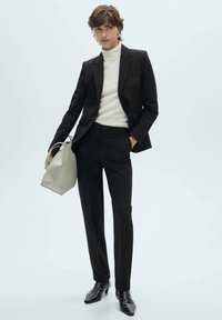 Abito scuro su misura con vestibilità slim, dolcevita bianco e scarpe nere. Tiene in mano una borsa grigia chiara. Tessuto liscio e design minimalista.