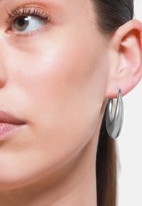 Pendientes de aro de metal plateado con un diseño suave y curvado. Los pendientes tienen un acabado brillante y presentan sutiles acentos cerca del cierre.
