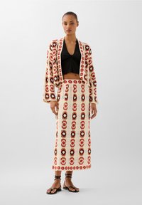 Pletený dvoudílný outfit složený z krátkého cardigan a dlouhé sukně. Krémový základ s červenými a černými květinovými vzory. Celkový vzhled doplňují ploché sandály.