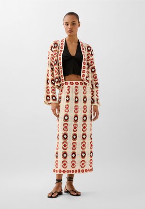 Completo due pezzi in maglia composto da un cardigan corto e una gonna lunga. Base crema con motivi floreali rossi e neri. Sandali piatti completano il look.
