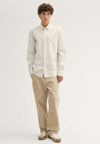 Helles beigefarbenes Hemd mit Knöpfen und strukturiertem Rautenmuster, kombiniert mit lockeren, beigen Cargo-Hosen und weißen Sneakers.