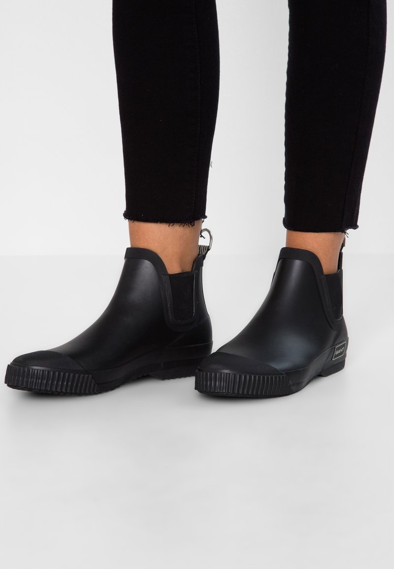 Zwarte rubberen enkelboots hebben een gladde afwerking, elastische zijkanten, een contrasterende textuurzool en een achterste trekkoortje voor eenvoudig aan- en uittrekken.
