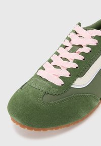 Grønn suedesneaker med rosa teksturerte snører, hvit side stripe og gummisåle. Har avrundet tå og sømdetaljer.