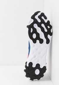 Skor med en texturerad svart gummisula som har cirkulära mönster och en vit mellansula, kompletterad med en blå ovandel och röda accenter.