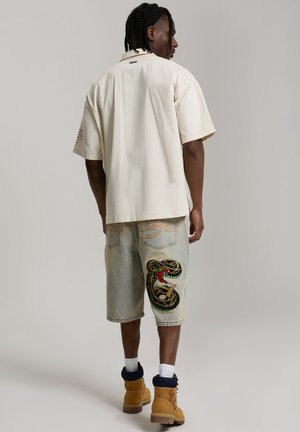 Uomo che indossa una camicia larga crema a maniche corte, pantaloncini di jeans con un grande disegno colorato di serpente, calzini bianchi e stivali da lavoro beige, visto di spalle.