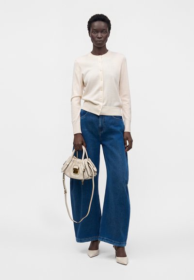 Femme en cardigan crème boutonné, jean bleu à jambes larges, escarpins crème pointus, tenant un petit sac à main crème avec une fermeture dorée.