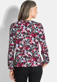 Frau mit langen grauen Haaren, die eine langärmelige Bluse mit roten, schwarzen, blauen und weißen geometrischen Mustern trägt, kombiniert mit schwarzen Hosen.