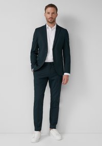 Traje ajustado en verde azulado oscuro con una camisa blanca, presenta un cierre de un solo botón y dos bolsillos frontales. Combinado con zapatillas blancas.